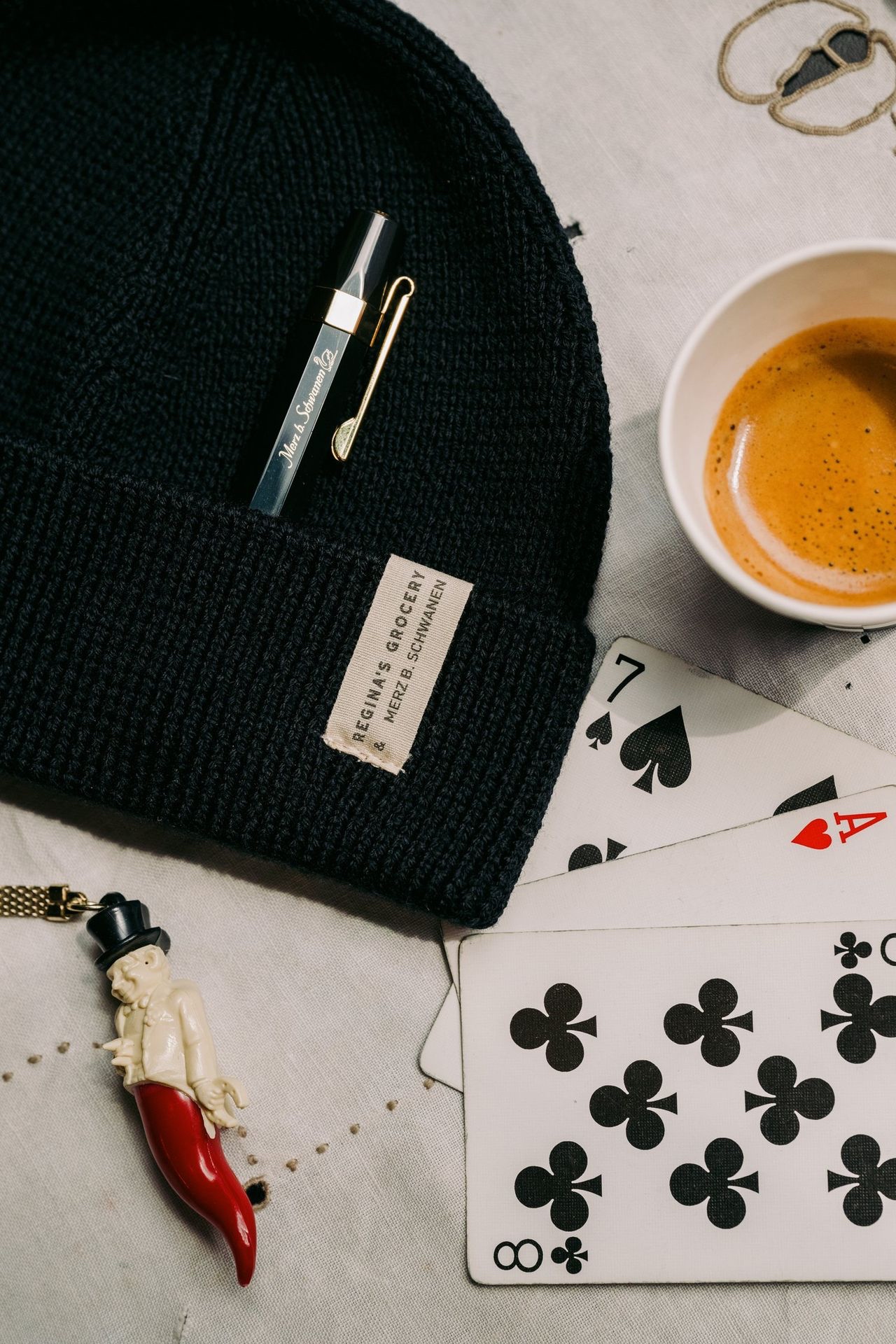 Black beanie and metal pencil next to coffee and playing cards