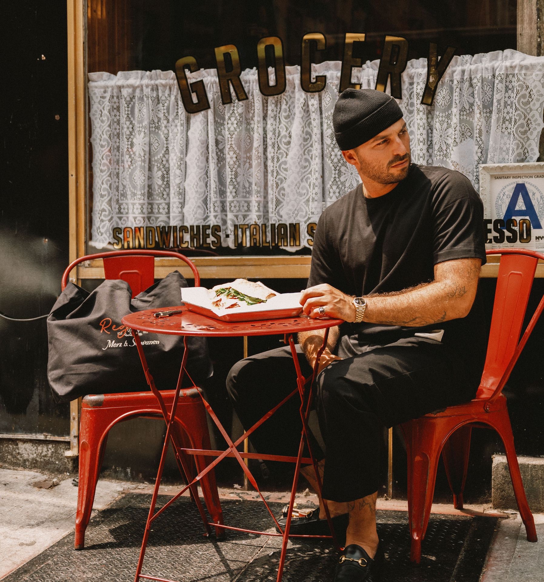 Man in black T-shirt, chino trousers and short beanie eating a sandwich