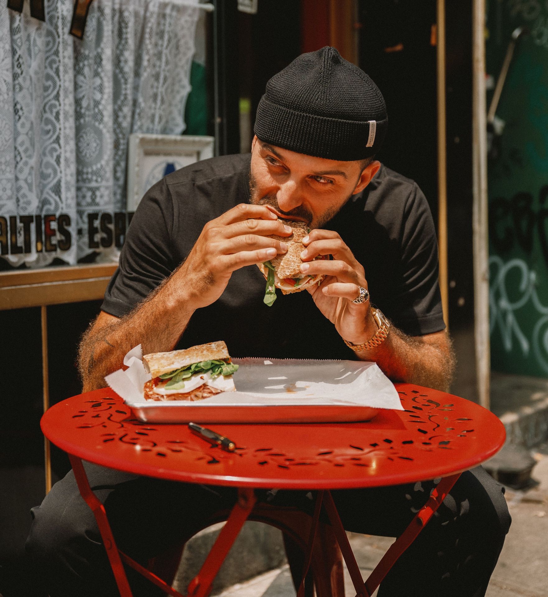 Man with black T-shirt and short beanie eating a sandwich
