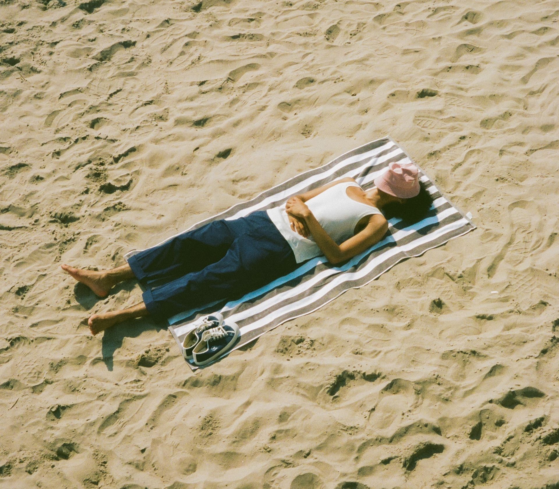 Man in blue chino trousers and white tank top laying at the beach with bucket hat covering his face