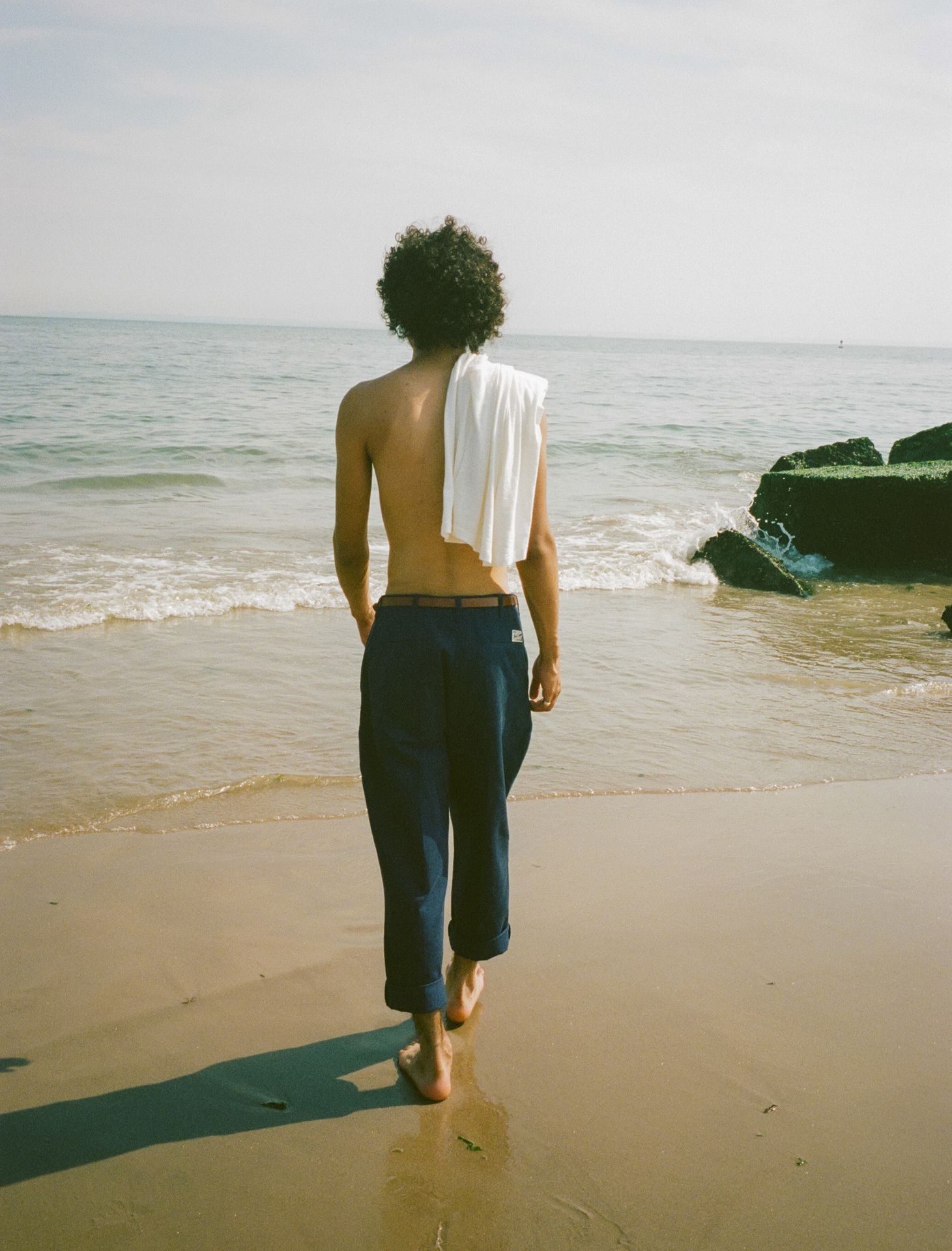 Man at the beach wearing blue chino trousers with white tanktop over his shoulders
