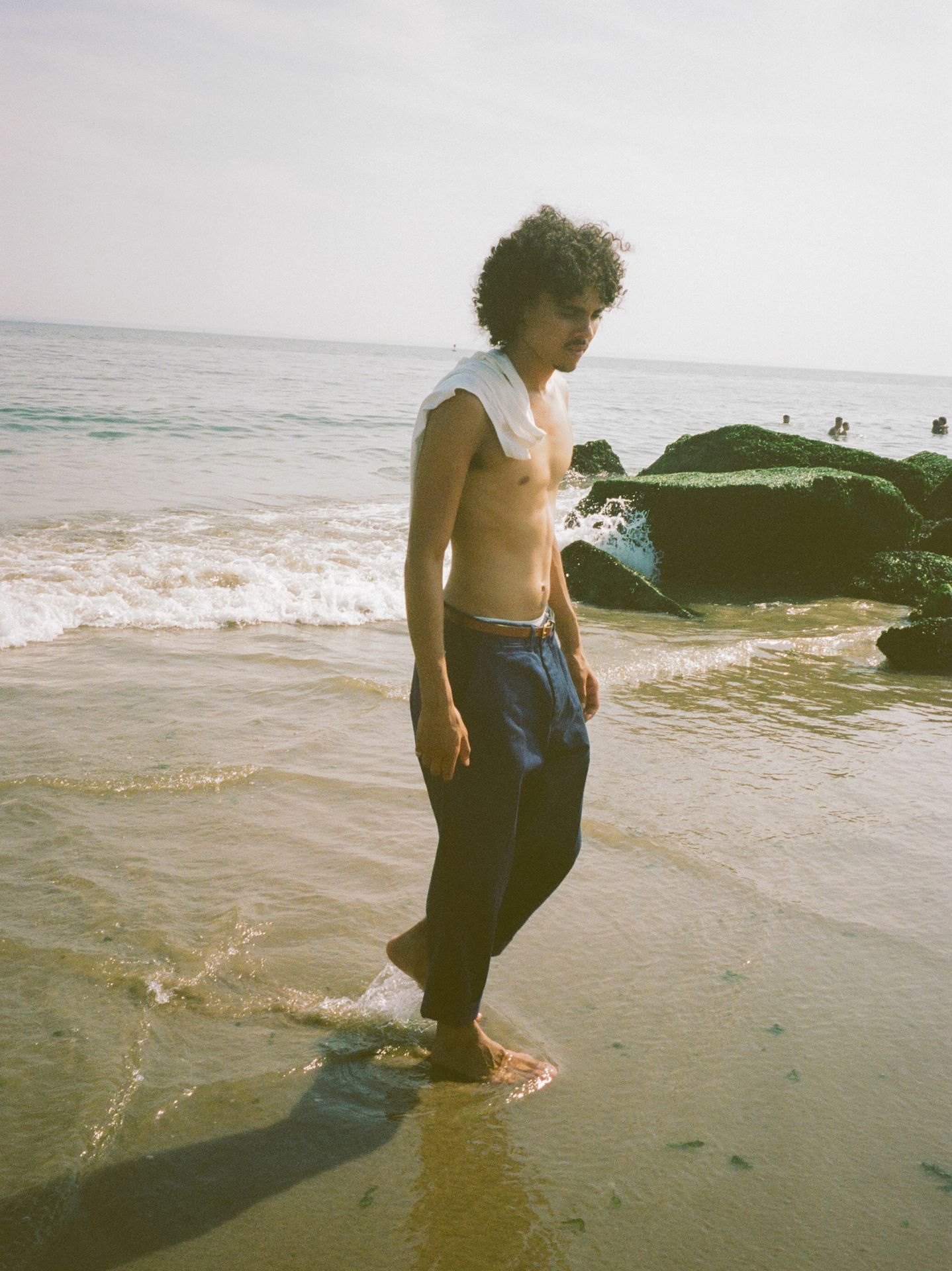 Man at the beach wearing blue chino trousers with white tanktop over his shoulders